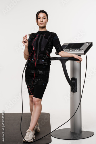 Foto Fit girl in EMS suit with contactor in hand that uses electrical impulses to stimulate muscles on white background