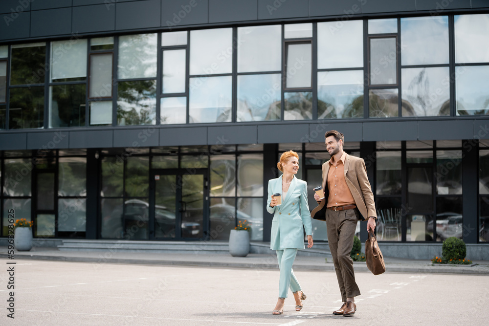 full length of trendy business partners with paper cups walking near modern building on urban street. full length of trendy business partners with paper cups walking near modern building on urban street.