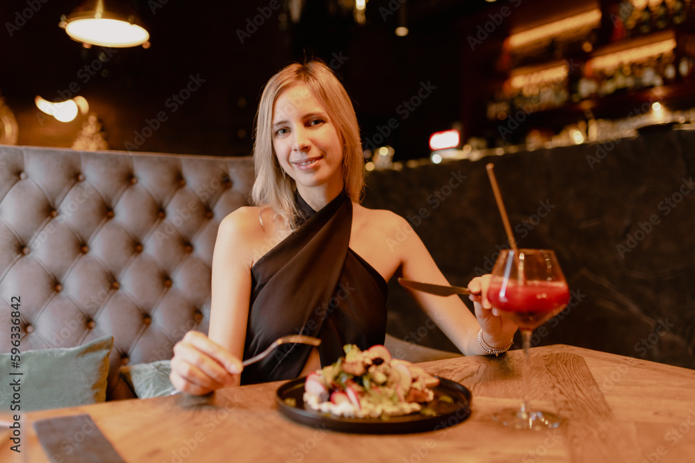 Portrait of young cheerful perfect woman sitting on brown sofa at wooden table near black plate with vegetable salad and glass with red cocktail, holding fork, knife in restaurant. Celebration, party.