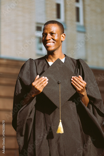 Happy african american student in mantle standing outdoors on sunny day and holding in hands graduation hat. Job search, graduate from university, employment, start in life, thoughts about future.