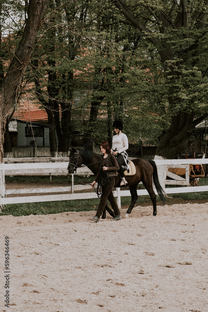 A woman instructor teaching girl how to ride a horse. Female rider practicing on a horseback learning equestrian sport. Active lifestyle and leisure activity concep A woman instructor teaching girl how to ride a horse. Female rider practicing on a horseback learning equestrian sport. Active lifestyle and leisure activity concep