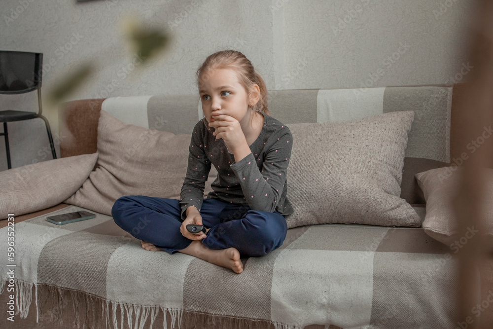 A cute little girl is sitting on the couch at home, using the remote control to switch channels on the TV. Watch informative and interesting children's programs or videos for children and teenagers A cute little girl is sitting on the couch at home, using the remote control to switch channels on the TV. Watch informative and interesting children's programs or videos for children and teenagers