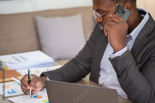 An African American businessman in a black suit uses a mobile phone to communicate with clients and a laptop to view company profits. to build confidence for customers to come and invest in the office