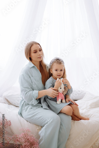 Happy young loving mother plays plush bunny with her daughter in the bedroom. Family in home clothes  blue pajamas and a dress.