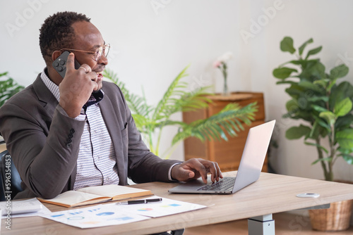 An African American businessman in a black suit uses a mobile phone to communicate with clients and a laptop to view company profits. to build confidence for customers to come and invest in the office