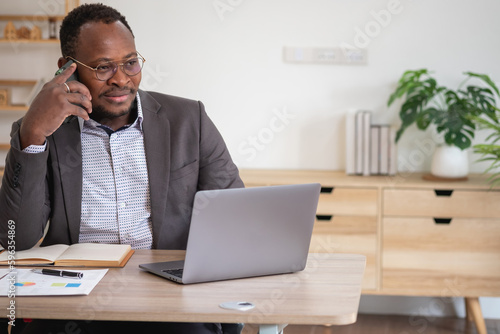 An African American businessman in a black suit uses a mobile phone to communicate with clients and a laptop to view company profits. to build confidence for customers to come and invest in the office