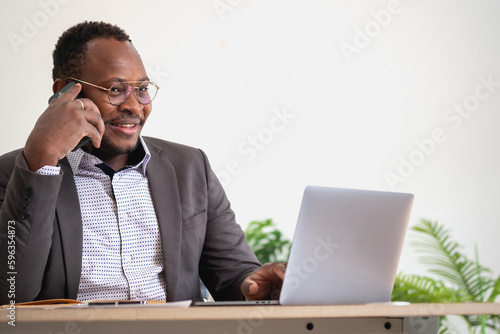 An African American businessman in a black suit uses a mobile phone to communicate with clients and a laptop to view company profits. to build confidence for customers to come and invest in the office
