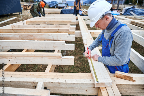 Man worker building wooden frame house on pile foundation. Carpenter using tape measure for measuring wooden planks and making marks with pencil. Carpentry concept.