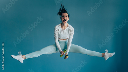 Young brunette American woman in sportswear jumps high holds dumbbells screams against blue wall. Healthy people. Fit American girl shouting out loud. Sport club, weight loss concept. Fitness class.