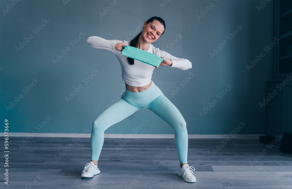 Brunette hispanic young woman in sportswear stretching rubber tape at fitness club looks at camera happily. Fit caucasian girl training at home. Fitness, sport and healthy people.