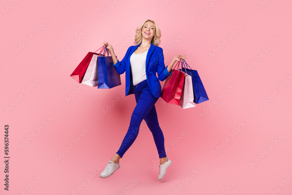 Full length photo of sweet cute lady wear blue jacket walking jumping high holding bargains isolated pink color background
