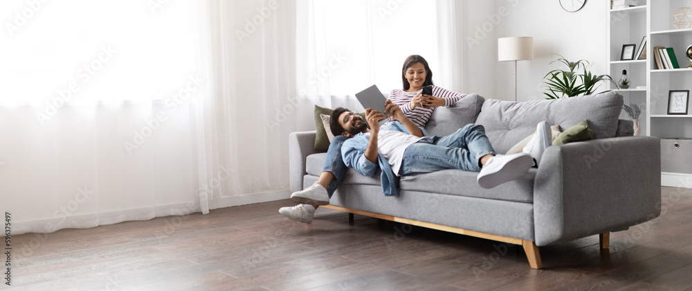 Relaxed indian spouses resting on sofa at home, using gadgets Relaxed indian spouses resting on sofa at home, using gadgets