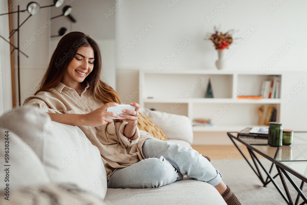 Cheerful millennial european female watching video on phone, surfing in internet in minimalist living room