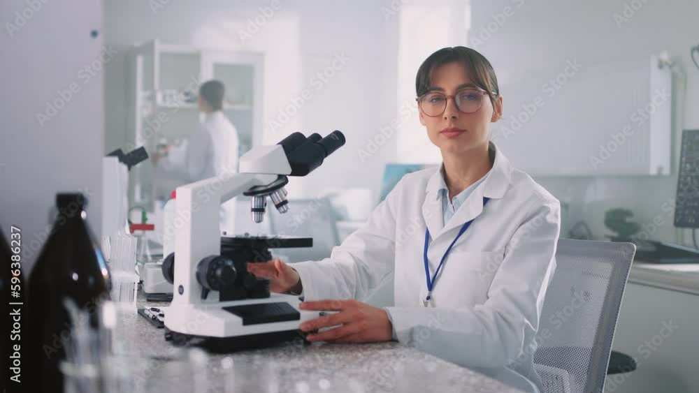 Horizontal shot of young Caucasian lab worker in white coat using microscope. Portrait of attractive brunette woman in glasses looking directly at camera indoors. Working day. Modern equipment in Horizontal shot of young Caucasian lab worker in white coat using microscope. Portrait of attractive brunette woman in glasses looking directly at camera indoors. Working day. Modern equipment in
