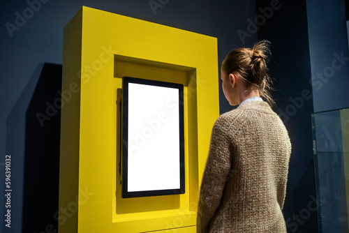 Woman looking at vertical blank digital interactive white display wall at exhibition or museum with futuristic interior. Mock up, future, copyspace, white screen, technology concept