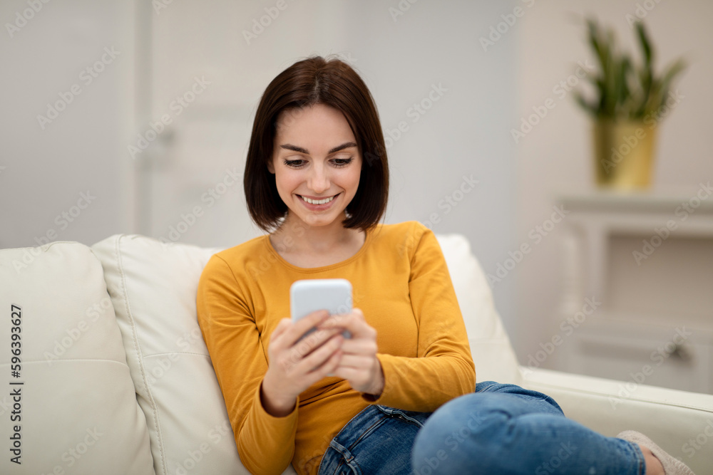 Cheerful young woman sitting on couch at home, using smartphone Cheerful young woman sitting on couch at home, using smartphone