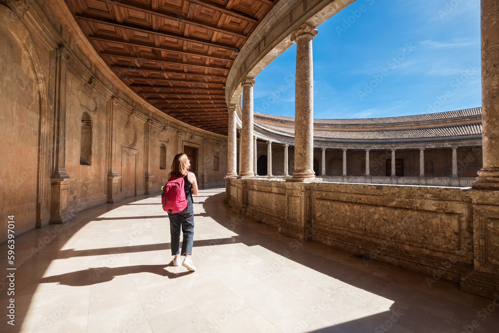 Young female tourist walking with backpack by the wide galleries with columns in the inner circular patio in Palace of Charles V in Alhambra complex in Granada, Andalusia, Spain. Young female tourist walking with backpack by the wide galleries with columns in the inner circular patio in Palace of Charles V in Alhambra complex in Granada, Andalusia, Spain.