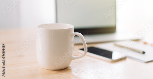 White mug without inscriptions opposite laptop and notepad with pen on wooden table.