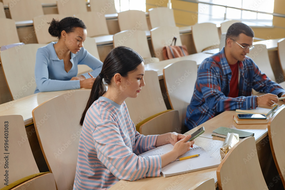 Group of students sitting at desk with books using mobile phones during lecture at university