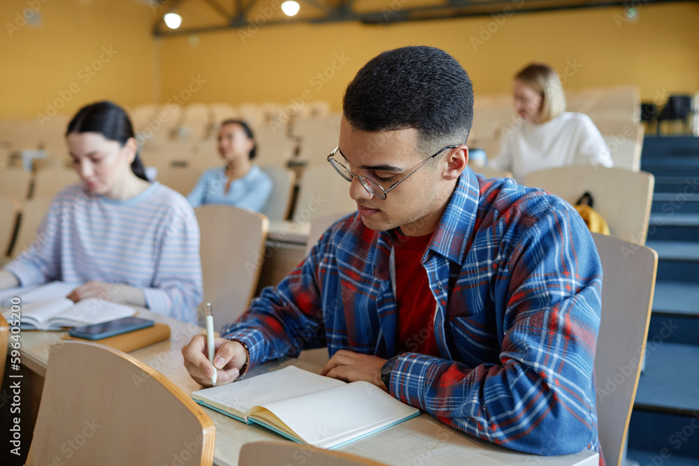 Teenage boy making notes in notebook while sitting at desk at lecture together with his classmates in background Teenage boy making notes in notebook while sitting at desk at lecture together with his classmates in background