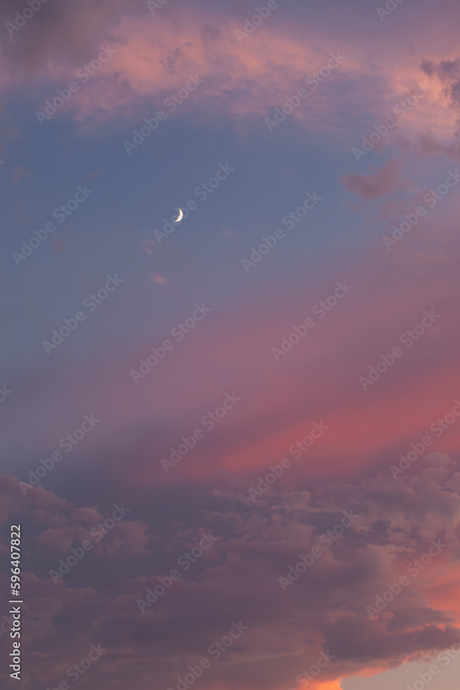 The orange pink sunset sky with a quarter moon. The quarter moon is visible in the blue sky. The clouds are pink to orange-grey. The orange pink sunset sky with a quarter moon. The quarter moon is visible in the blue sky. The clouds are pink to orange-grey.