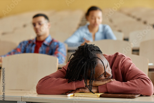 African American schoolboy lying on desk and sleeping during boring lecture with his classmates in background photo