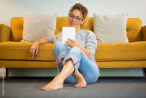 Easy relaxed lady at home reading and using tablet ereader ebook device alone witting on the ground against the sofa. Relaxation and independent lifestyle female people. Young adult lady in indoor photo