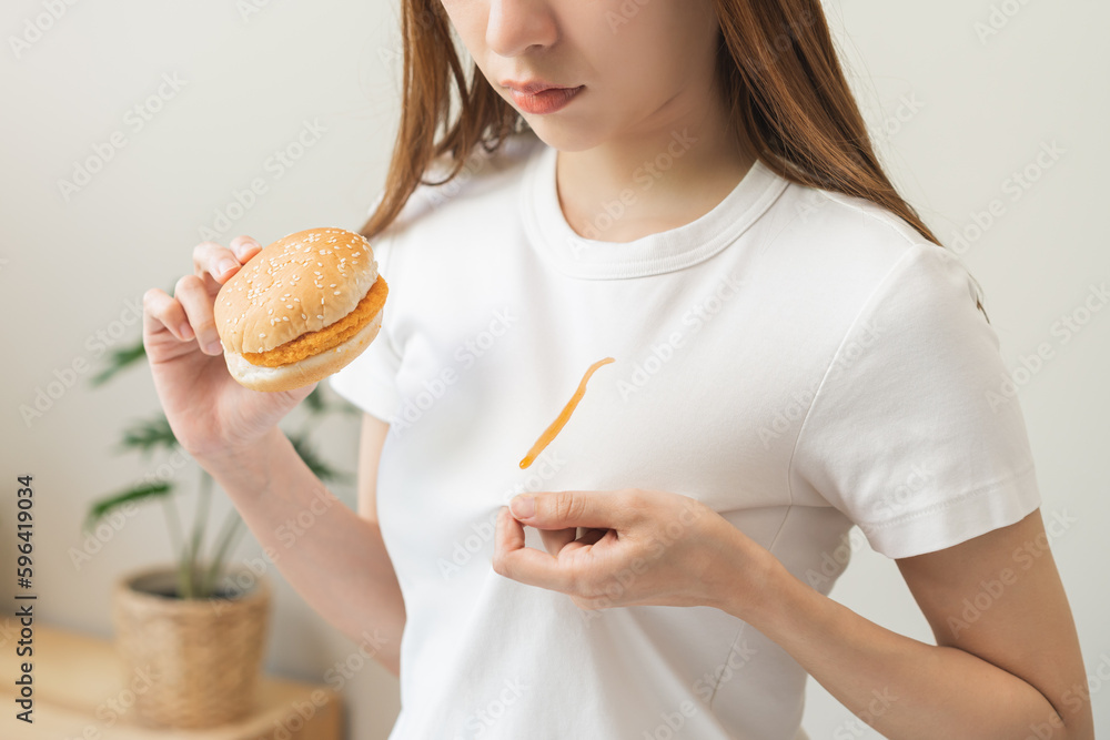 Cloth stain, disappointment asian young woman, girl eat food hand hold hamburger, burger show making tomato sauce smudged spilling, drop on white t-shirt, spot dirty or smudge on clothes, dirt stains. Cloth stain, disappointment asian young woman, girl eat food hand hold hamburger, burger show making tomato sauce smudged spilling, drop on white t-shirt, spot dirty or smudge on clothes, dirt stains.