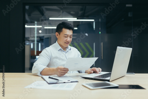 Office work. A young Asian man is working on a laptop and documents while sitting at a table.