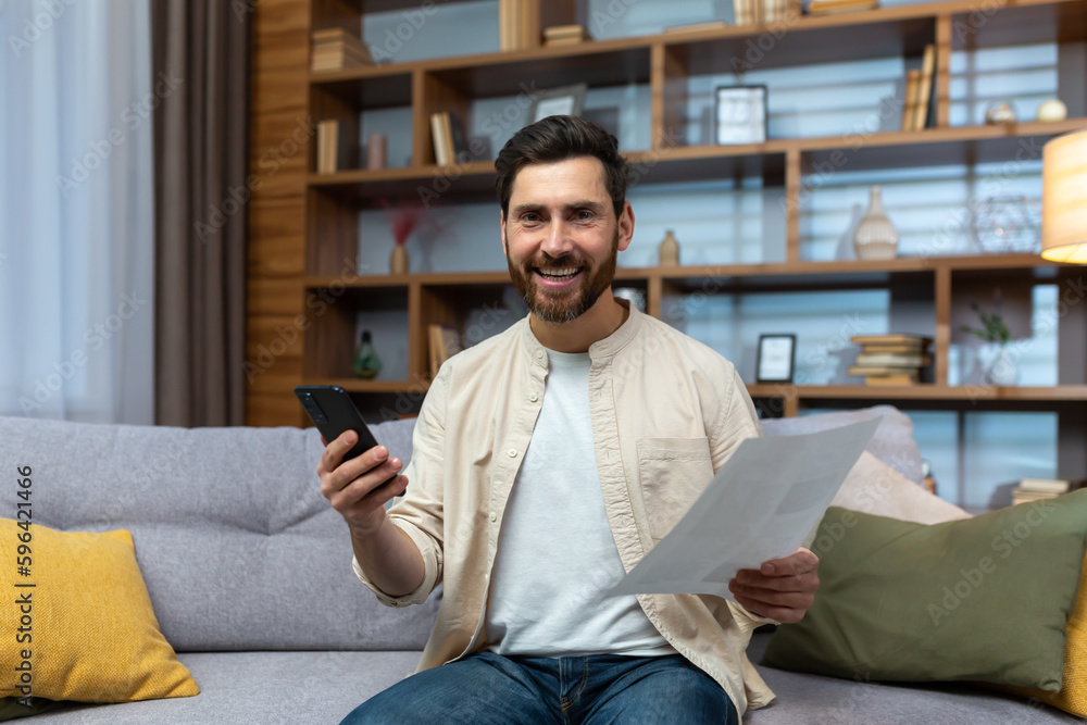 Portrait of a happy young man sitting at home on the couch. a smiling man looks at the camera, holds a phone and documents, papers in his hands.