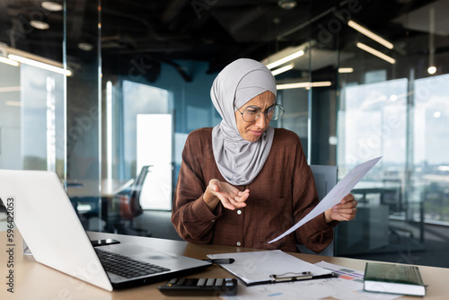 Financial problems, broken contracts, agreements. Confused young Muslim hijabi businesswoman sitting in the office, working on a laptop and holding documents, graphs, a project in her hands.