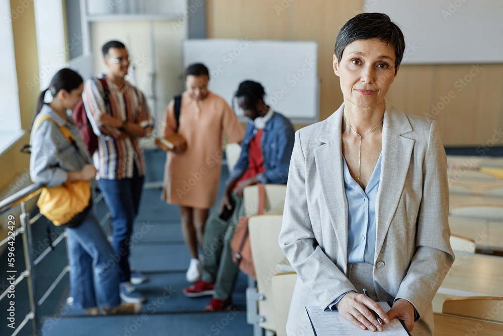 Portrait of mature teacher in elegant suit looking at camera while standing in lecture hall after lecture with students in background Portrait of mature teacher in elegant suit looking at camera while standing in lecture hall after lecture with students in background