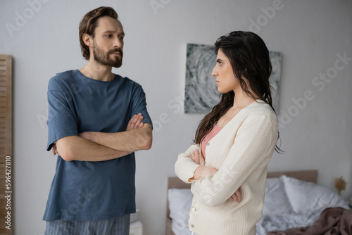 Offended woman crossing arms and looking at bearded boyfriend in pajama in bedroom.