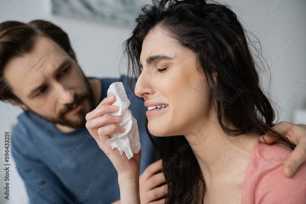Blurred man calming down displeased and crying girlfriend with napkin at home. Blurred man calming down displeased and crying girlfriend with napkin at home.