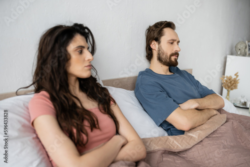 Dissatisfied man crossing arms while sitting near blurred girlfriend on bed at home.