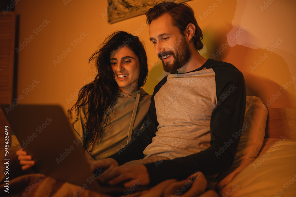 Carefree couple using blurred laptop on bed at night. Carefree couple using blurred laptop on bed at night.