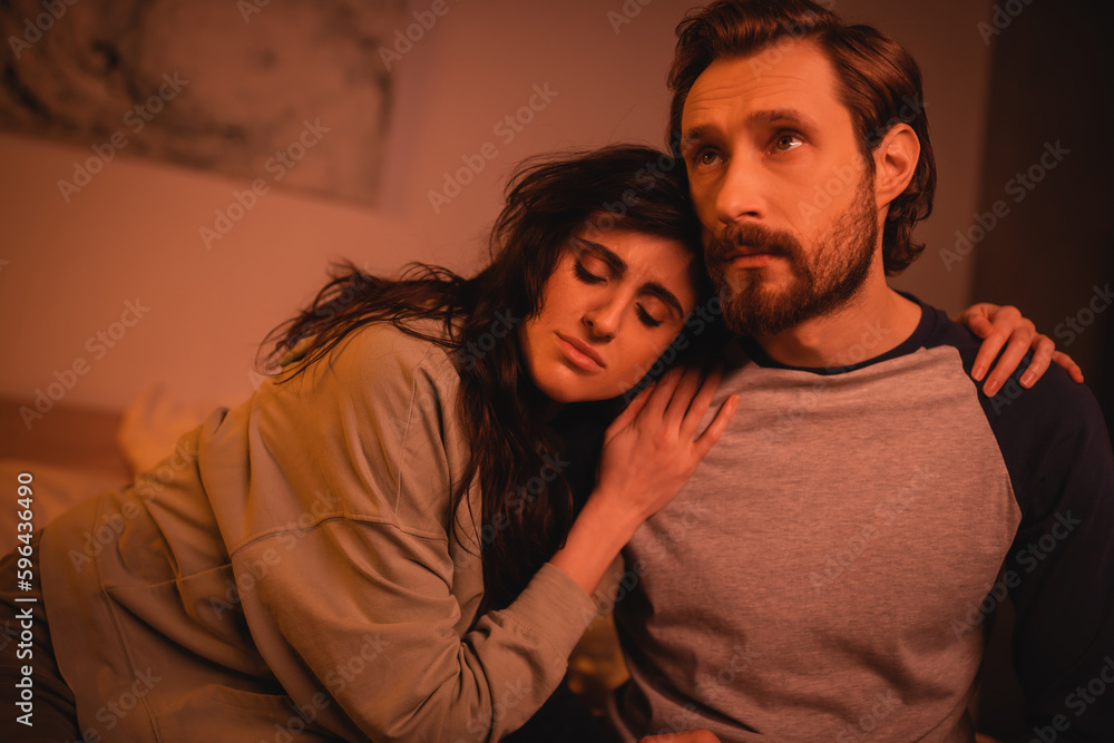 Brunette woman hugging sad boyfriend in bedroom at night.