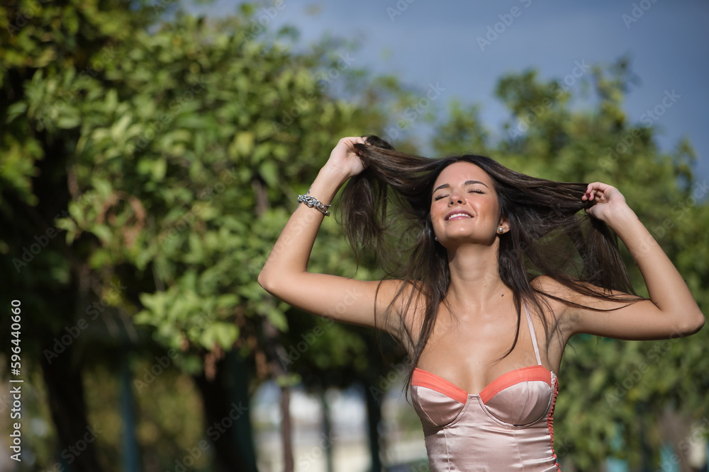 Young and beautiful woman with straight brown hair, orange top, with her eyes closed and pulling her hair with both hands smiling and happy. Concept fashion, beauty, trend, relax, millennial.