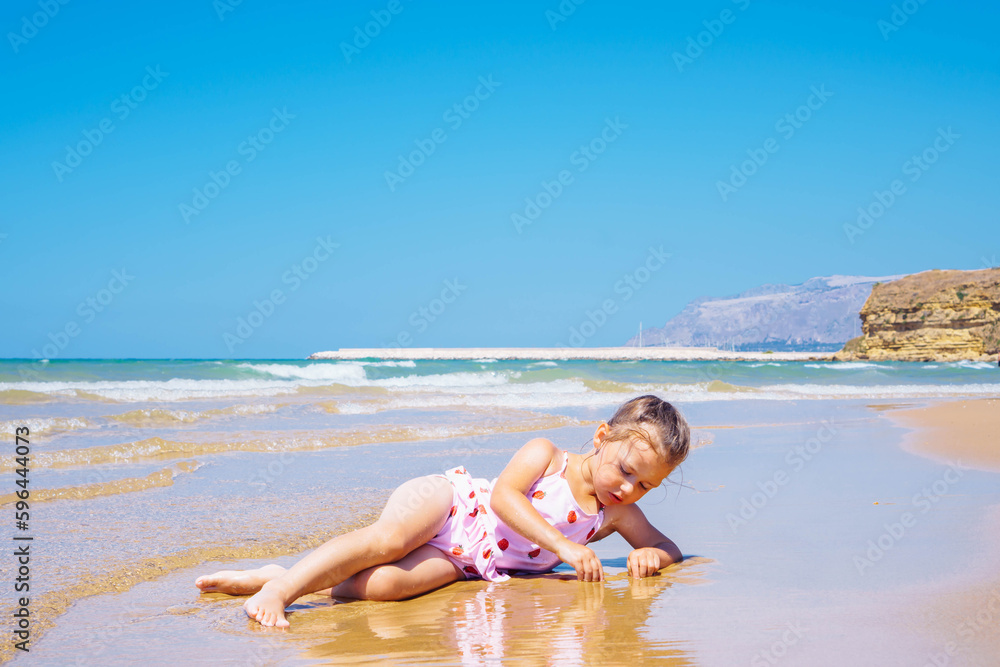 Cute little girl in pink swimsuit with strawberries lying in the sand and  playing with water