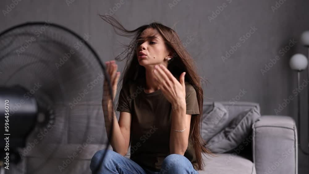 Young woman standing in front of fan while showing very hot temperature inside home Young woman standing in front of fan while showing very hot temperature inside home