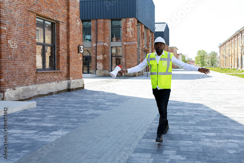 African american builder with megaphone in hand dancing at construction site. Joyful foreman in vest and hardhat enjoying with successful work. Achievement and people concept.