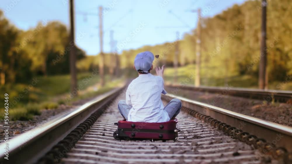 Solitude on the Rails: Outdoor Shot of a Melancholic Boy in a Cap Sitting on a Suitcase Solitude on the Rails: Outdoor Shot of a Melancholic Boy in a Cap Sitting on a Suitcase