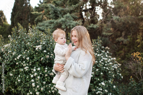 Happy smiling young mother holding baby 1 year old wearing stylish casual knitted clothes standing over blooming bushes with flowers in park outdoor. Motherhood. Maternity.