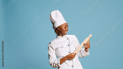 Smiling positive cook playing with rolling pin and singing on camera, having fun with cooking utensils. Young female chef holding wooden paddle, gastronomy tools over blue background.