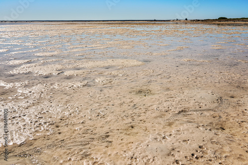 Sun shines to shallow sea during low tide, wet sand visible, sandy shore in distance
