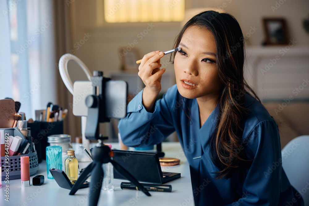 Fototapeta premium Young Asian woman looking herself on smart phone's screen while applying make up at home.