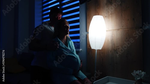 Husband caressing his pregnant wife sitting on the bed. Woman prepares for childbirth in hospital. Dark room backdrop. photo