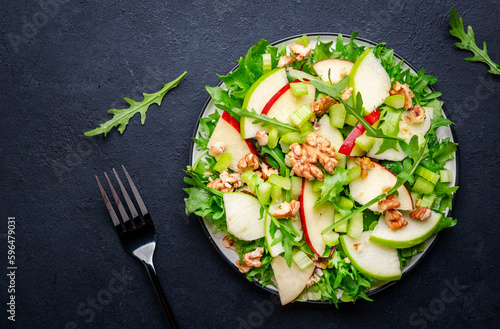Waldorf salad with red and green apple, celery, lettuce, arugula and walnuts on plate, black table background, top view