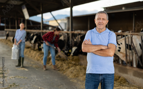 Portrait of confident mature male farmer posing in cowshed at dairy farm on summer day