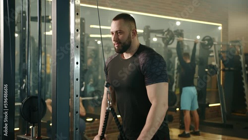 Slow motion medium shot of athletic young man focused on training on vertical row machine in gym. Man and woman exercising with barbells in the background. High quality 4k footage